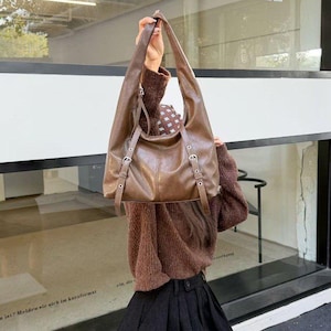 Puede incluir: Un bolso de hombro de cuero marr&oacute;n con correas largas y hebillas plateadas. El bolso lo sostiene una persona. El bolso tiene un dise&ntilde;o holgado y una forma rectangular. El fondo es una ventana.