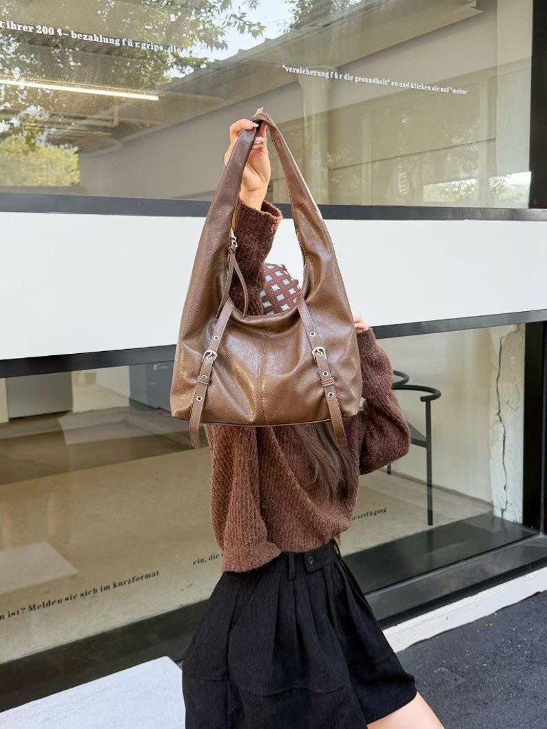 Puede incluir: Un bolso de hombro de cuero marr&oacute;n con correas largas y hebillas plateadas. El bolso lo sostiene una persona. El bolso tiene un dise&ntilde;o holgado y una forma rectangular. El fondo es una ventana.