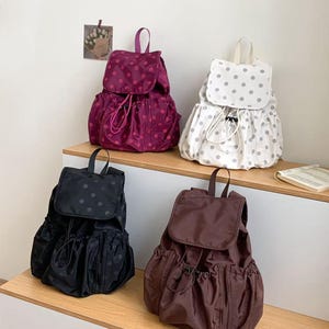 Può includere: Quattro zaini con coulisse in vari colori: bordeaux a pois rosa, bianco a pois grigi, nero a pois neri e marrone. Ogni zaino ha una chiusura a patta e un manico superiore. Gli zaini sono esposti su una mensola di legno.