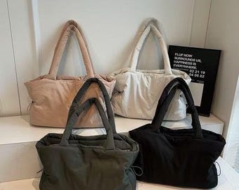 Könnte beinhalten: Vier Tragetaschen in den Farben Rosé, Off-White, Olivgrün und Schwarz. Jede Tasche hat ein gepufftes Design und passende Griffe. Im Hintergrund befindet sich ein schwarz-weißes Schild mit Text.