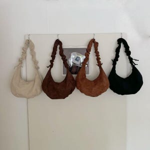 May include: Four suede shoulder bags in cream, brown, tan, and black hang from a white wall. Each bag has a crescent shape with a gathered strap. The bags are a neutral color palette.