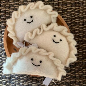 May include: Three white felt dumplings with ruffled edges and smiling faces sit in a small wooden bowl. The dumplings have black stitched smiles. The background is a textured, dark brown and grey surface.