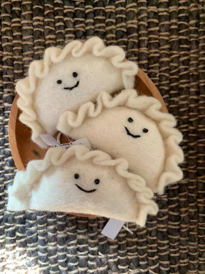 May include: Three white felt dumplings with ruffled edges and smiling faces sit in a small wooden bowl. The dumplings have black stitched smiles. The background is a textured, dark brown and grey surface.