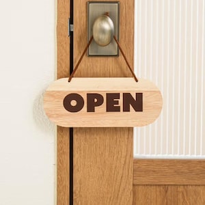 以下が含まれることがあります： 木製の「OPEN」サインが木のドアに吊り下げられています。サインは楕円形で、角が丸く、茶色の文字が書かれています。ドアには金属製の取っ手と、縦線が入ったガラスパネルが付いています。