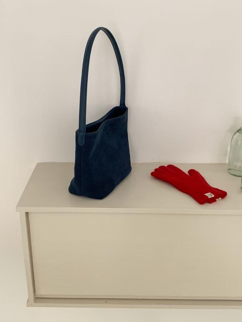 May include: A navy blue tote bag with a long handle and a pair of red gloves. The bag is made of a soft material and has a simple design. The gloves are made of a warm material.
