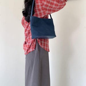 May include: A navy blue shoulder bag with a rectangular shape and a long strap. The bag is made of a soft material and has a simple design. The person is wearing a red plaid shirt and a gray skirt.