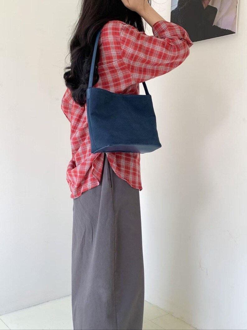 May include: A navy blue shoulder bag with a rectangular shape and a long strap. The bag is made of a soft material and has a simple design. The person is wearing a red plaid shirt and a gray skirt.