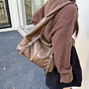 Puede incluir: Un bolso de hombro de cuero marr&oacute;n con hebillas y correas plateadas. El bolso tiene una forma rectangular y una correa larga para llevarlo al hombro. El bolso est&aacute; hecho de cuero liso y brillante.