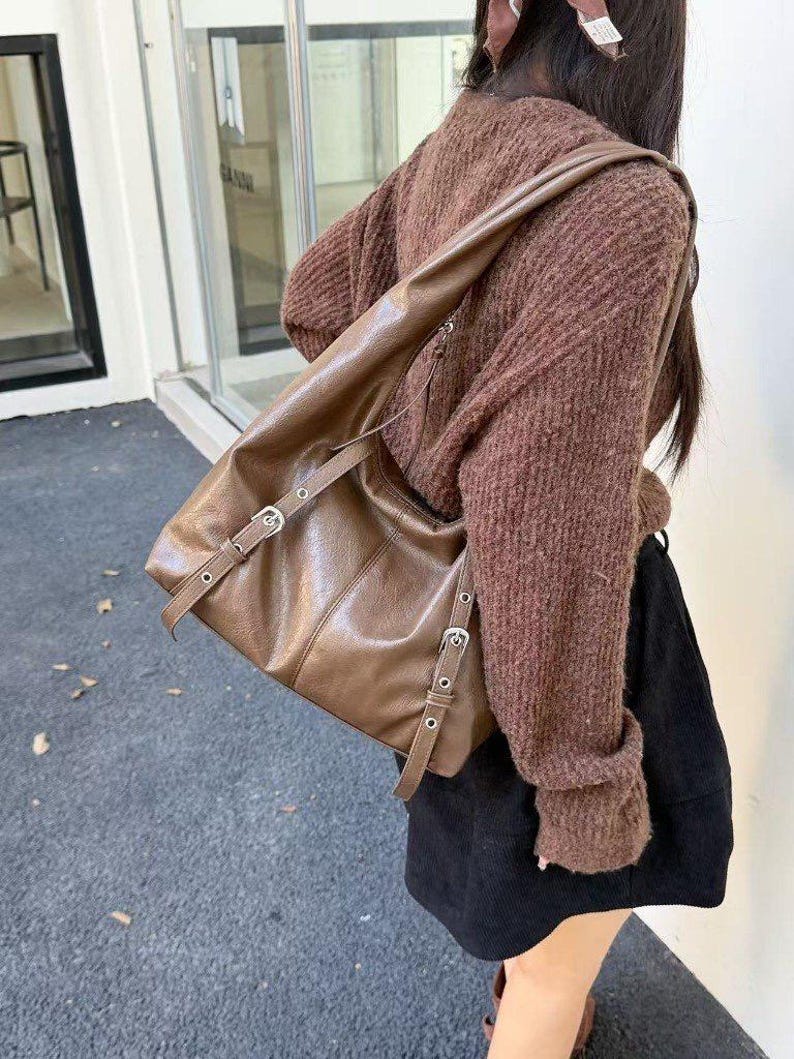 Puede incluir: Un bolso de hombro de cuero marr&oacute;n con hebillas y correas plateadas. El bolso tiene una forma rectangular y una correa larga para llevarlo al hombro. El bolso est&aacute; hecho de cuero liso y brillante.
