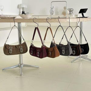 May include: A collection of six suede shoulder bags in various colors, including brown, burgundy, and black, are displayed hanging from a wooden table. Each bag features a flap closure with a silver buckle and a shoulder strap.