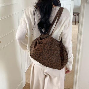 Puede incluir: Un bolso de hombro marr&oacute;n con estampado de leopardo y forma redondeada. El bolso tiene una correa larga y un peque&ntilde;o bolsillo. La persona lleva un su&eacute;ter blanco, pantalones beiges y un gorro marr&oacute;n.