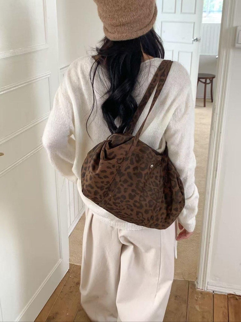 Puede incluir: Un bolso de hombro marr&oacute;n con estampado de leopardo y forma redondeada. El bolso tiene una correa larga y un peque&ntilde;o bolsillo. La persona lleva un su&eacute;ter blanco, pantalones beiges y un gorro marr&oacute;n.
