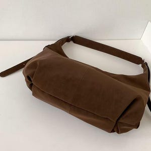 May include: A brown shoulder bag with a long strap. The bag is made of a soft, suede-like material. The bag has a rectangular shape with a curved top and a long, thin strap. The bag is sitting on a white surface.