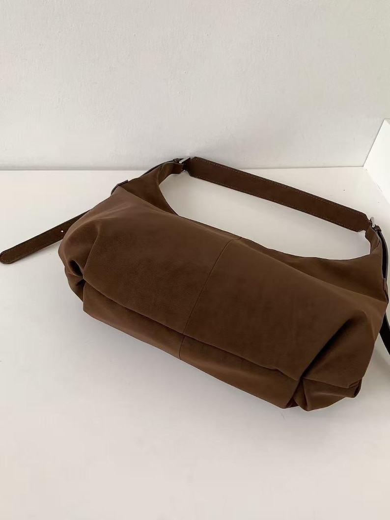 May include: A brown shoulder bag with a long strap. The bag is made of a soft, suede-like material. The bag has a rectangular shape with a curved top and a long, thin strap. The bag is sitting on a white surface.