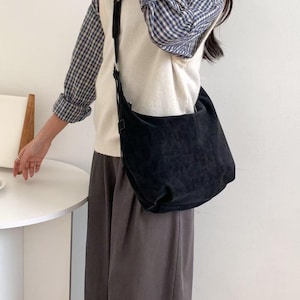 May include: A black suede shoulder bag with a long strap. The bag is worn over a white sweater vest and a blue and white plaid shirt. The person is wearing gray pants.