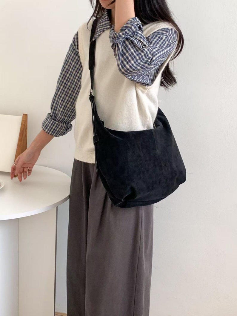May include: A black suede shoulder bag with a long strap. The bag is worn over a white sweater vest and a blue and white plaid shirt. The person is wearing gray pants.