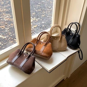 Może przedstawiać: Cztery torebki w różnych kolorach, w tym brązowym, beżowym i czarnym, są wystawione na białym parapecie. Torby mają górne uchwyty i strukturalne kształty, z widocznymi szwami i detalami okuć. Torby wydają się być wykonane z zamszu i skóry.