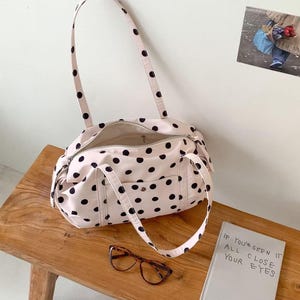 Puede incluir: Un bolso tote rosa claro con lunares negros, cierre de cremallera y dos asas. El bolso est&aacute; abierto, revelando el interior. Unas gafas carey y un libro con el texto "IF YOU'VE SEEN IT ALL, CLOSE YOUR EYES" est&aacute;n sobre un banco de madera.