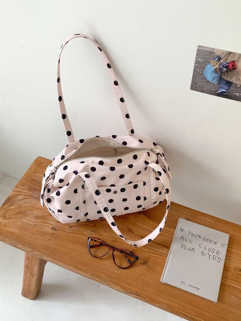 Puede incluir: Un bolso tote rosa claro con lunares negros, cierre de cremallera y dos asas. El bolso est&aacute; abierto, revelando el interior. Unas gafas carey y un libro con el texto "IF YOU'VE SEEN IT ALL, CLOSE YOUR EYES" est&aacute;n sobre un banco de madera.