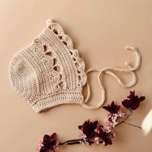 Puede incluir: Un gorro de punto de color beige claro con un delicado patrón de encaje. El gorro tiene una cinta que se puede atar debajo de la barbilla.
