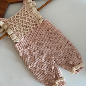 May include: A handmade, light pink crochet baby romper with cream-colored accents. The romper features a textured stitch pattern, ruffled edges, and a lattice design on the bodice. The garment is displayed on a wooden hanger.