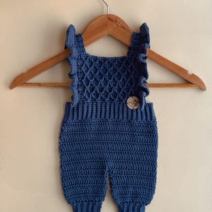 May include: A handmade, blue crochet baby romper with a textured bodice and ruffled straps. The romper is displayed on a wooden hanger. A small wooden button secures the front. The garment is a soft, comfortable material.