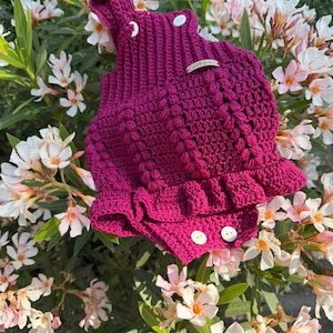 May include: A handmade magenta crochet romper with white buttons and a ruffled hem. The romper features a textured stitch pattern and is designed for a baby or toddler. The garment is displayed outdoors among green leaves and white and pink flowers.
