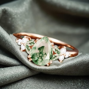 May include: A rose gold ring with a pear-shaped green agate gemstone surrounded by smaller clear gemstones.