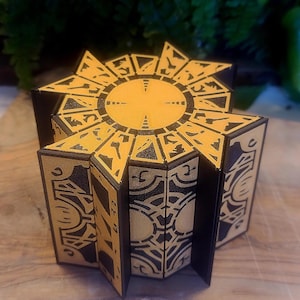 May include: A decorative puzzle box with a geometric design. The box is primarily black and gold, with intricate patterns and a starburst-like top. The box is open, revealing its interior.