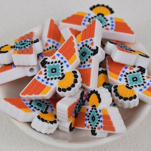 Op de afbeelding: Een verzameling witte siliconen kralen met een kleurrijk geometrisch ontwerp in oranje, geel, blauw en turquoise. De kralen hebben een gestileerd vogelontwerp.