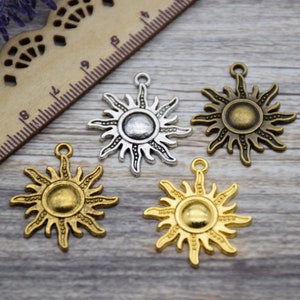 May include: Four sun charm pendants in silver and gold tones. The charms are in the shape of a sun with a circular centre and rays extending outwards. The charms are on a wooden surface.