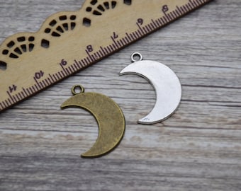 May include: Two crescent moon charms, one bronze-toned and one silver-toned, with small loops for hanging. A wooden ruler is in the background. These charms are suitable for jewelry making or craft projects.