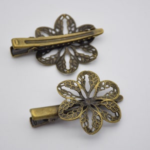 Könnte beinhalten: Zwei antike goldfarbene Haarklammern aus Metall mit einem zarten Blumenmuster. Jede Klammer zeigt eine Blume mit filigranen Ausschnitten und eine einfache, gerade Stangenklammer.