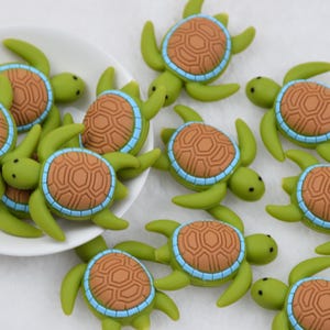 May include: A collection of green turtle-shaped embellishments with brown shell details and blue trim. Some turtles are in a white bowl, while others are scattered on a white surface. These decorative elements are suitable for crafts and DIY projects.