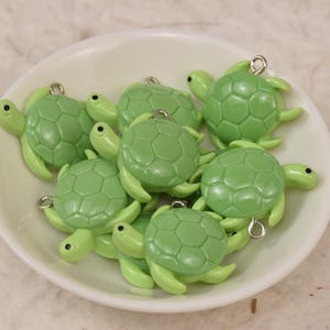 May include: A collection of small, light green turtle charms with silver-colored loops for attaching to jewelry or other crafts. The turtles are in a white bowl, with a light-colored background.