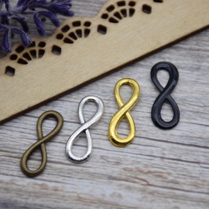 Pode incluir: Quatro pingentes com símbolo de infinito em diferentes acabamentos: dourado, prateado, bronze e preto. Os pingentes estão sobre uma superfície de madeira.