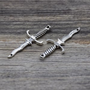 May include: Two silver-toned sword charms with intricate details. The swords have a pointed tip and a curved handle.