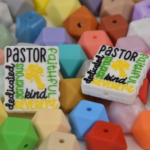 Pode incluir: Uma coleção de contas de silicone geométricas coloridas em várias tonalidades pastel e vibrantes. Duas contas retangulares brancas apresentam a palavra "PASTOR" e outras palavras como "dedicado" e "gentil" com um gráfico de cruz amarela.