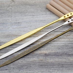 Op de afbeelding: Drie metalen gereedschappen met verschillende afwerkingen: goud, zilver en brons. Elk gereedschap heeft een puntig uiteinde en een afgeronde handgreep met gaten.