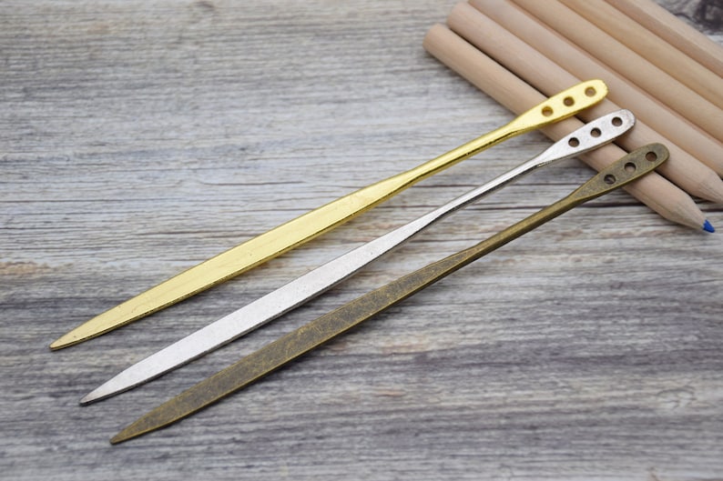 Op de afbeelding: Drie metalen gereedschappen met verschillende afwerkingen: goud, zilver en brons. Elk gereedschap heeft een puntig uiteinde en een afgeronde handgreep met gaten.