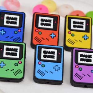 Może przedstawiać: Kolekcja kolorowych, miniaturowych konsol do gier przenośnych. Każda konsola ma czarną obwódkę, ekran z napisem "GAME OVER" i krzyżak. Kolory obejmują niebieski, czerwony, żółty, zielony, jasnoniebieski i fioletowy.