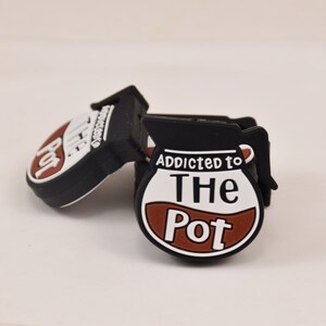 May include: Two black shoe charms with a coffee pot design. One charm is tilted, showing the text "Pot" in a white oval with a brown coffee stain. The other charm shows the text "Addicted to The Pot".