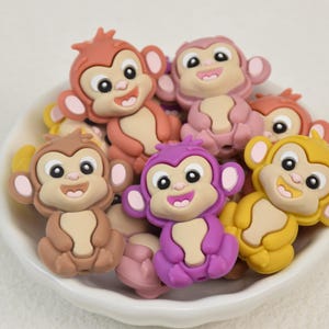 May include: A collection of colorful monkey-shaped embellishments in a white bowl. The monkeys are in shades of brown, pink, purple, and yellow, with smiling faces and large ears. These decorative pieces are ideal for crafts and DIY projects.
