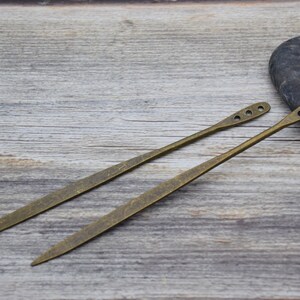 Op de afbeelding: Twee antieke goudkleurige metalen haarstokken met decoratieve gaten. De stokken liggen op een houten oppervlak.