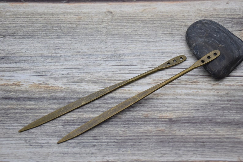 Op de afbeelding: Twee antieke goudkleurige metalen haarstokken met decoratieve gaten. De stokken liggen op een houten oppervlak.