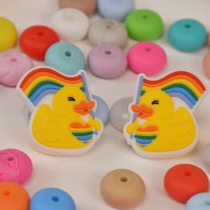 May include: Two yellow duck charms with rainbow flags, set amongst a variety of colourful beads. The charms have a white border and are smiling. The beads are in shades of pink, blue, green, yellow, and purple.
