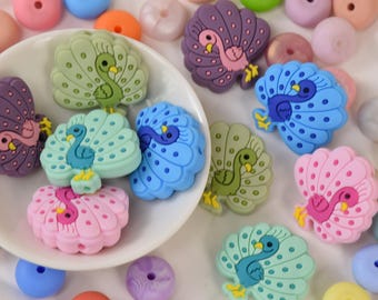 Könnte beinhalten: Eine Sammlung bunter Silikonperlen in Pfauenform in verschiedenen Farben wie Rosa, Blau, Grün und Lila. Die Perlen sind um eine weiße Schüssel herum angeordnet und von anderen bunten Perlen umgeben.