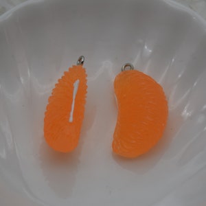 May include: Two orange, citrus-shaped charms with silver-colored loops for jewelry making. One charm is a segment, the other is a half-segment. The charms are set against a white background.