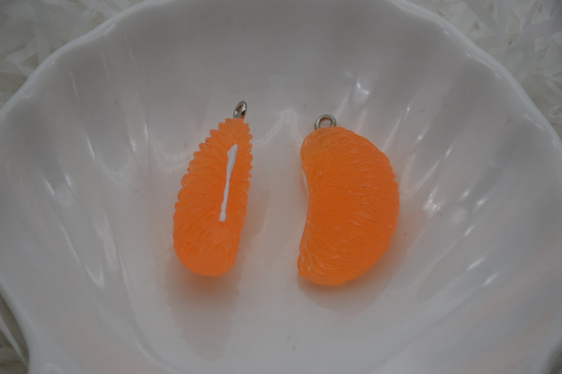 May include: Two orange, citrus-shaped charms with silver-colored loops for jewelry making. One charm is a segment, the other is a half-segment. The charms are set against a white background.
