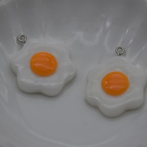 May include: Two white and orange fried egg charms with silver-colored loops for jewelry making. The egg yolks are bright orange, and the whites are a wavy, irregular shape. The charms are set against a white background.